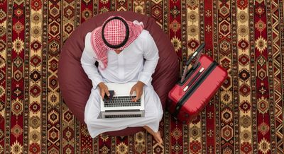 A Gulf man using a laptop with a red suitcase next to him