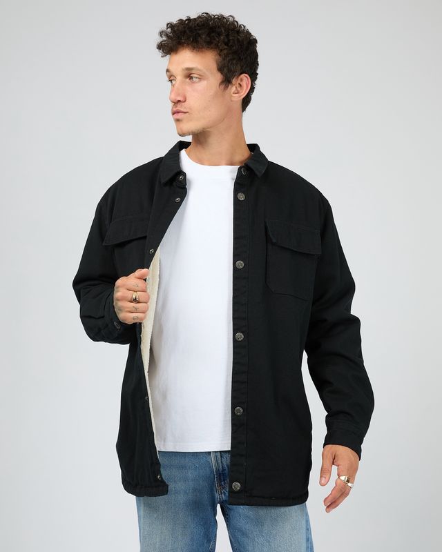 A male model wearing the Riot Sherpa Shacket in Black. a sherpa lined denim jacket with press stud buttons