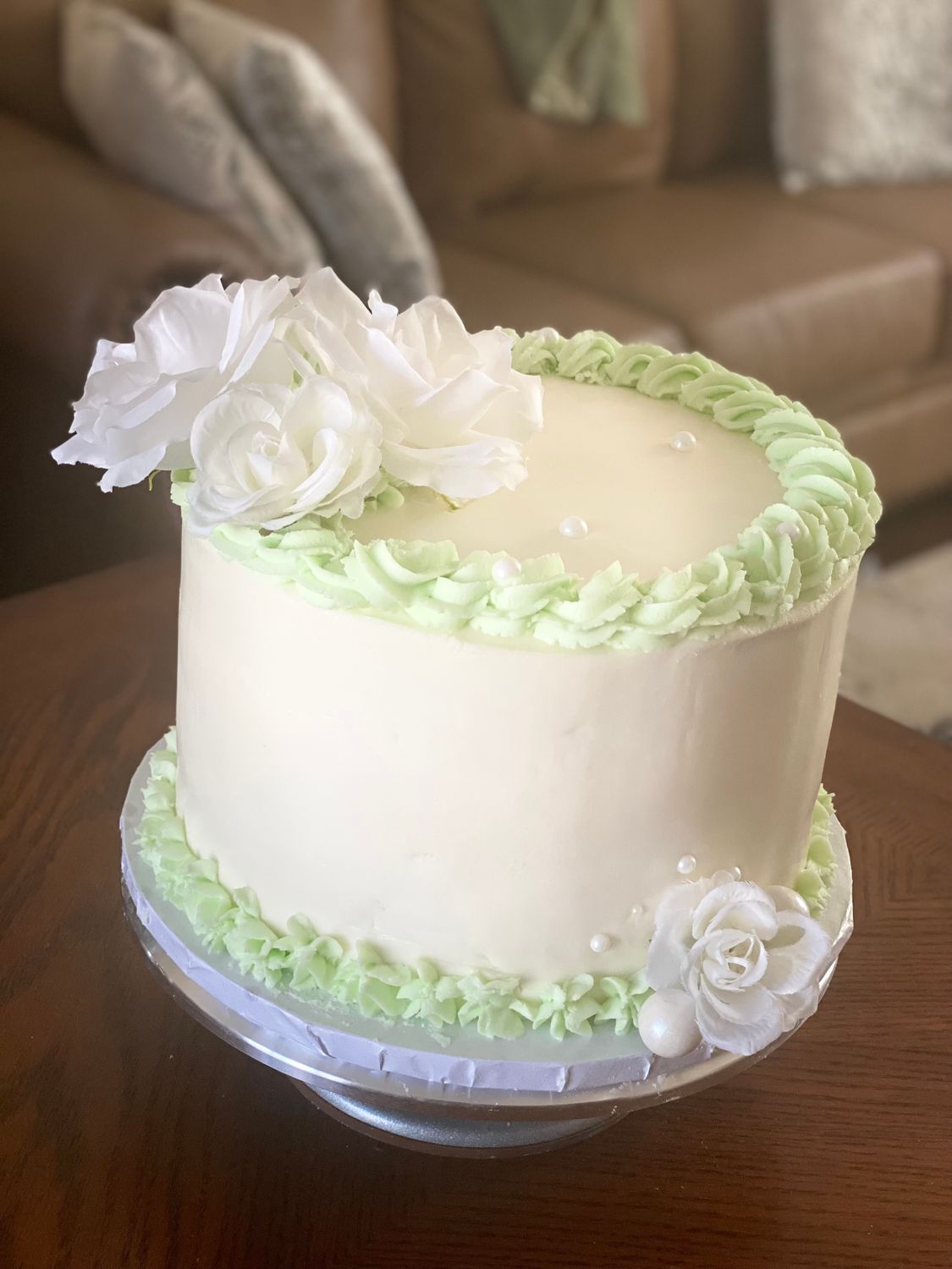 A beautiful 10-inch cake with a white base and sage green piping around the top and bottom edges with white flowers and white sugar pearls on the top corner and opposite the bottom corner of the cake. Perfect for special celebrations.