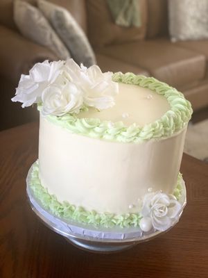 A beautiful 10-inch cake with a white base and sage green piping around the top and bottom edges with white flowers and white sugar pearls on the top corner and opposite the bottom corner of the cake. Perfect for special celebrations.