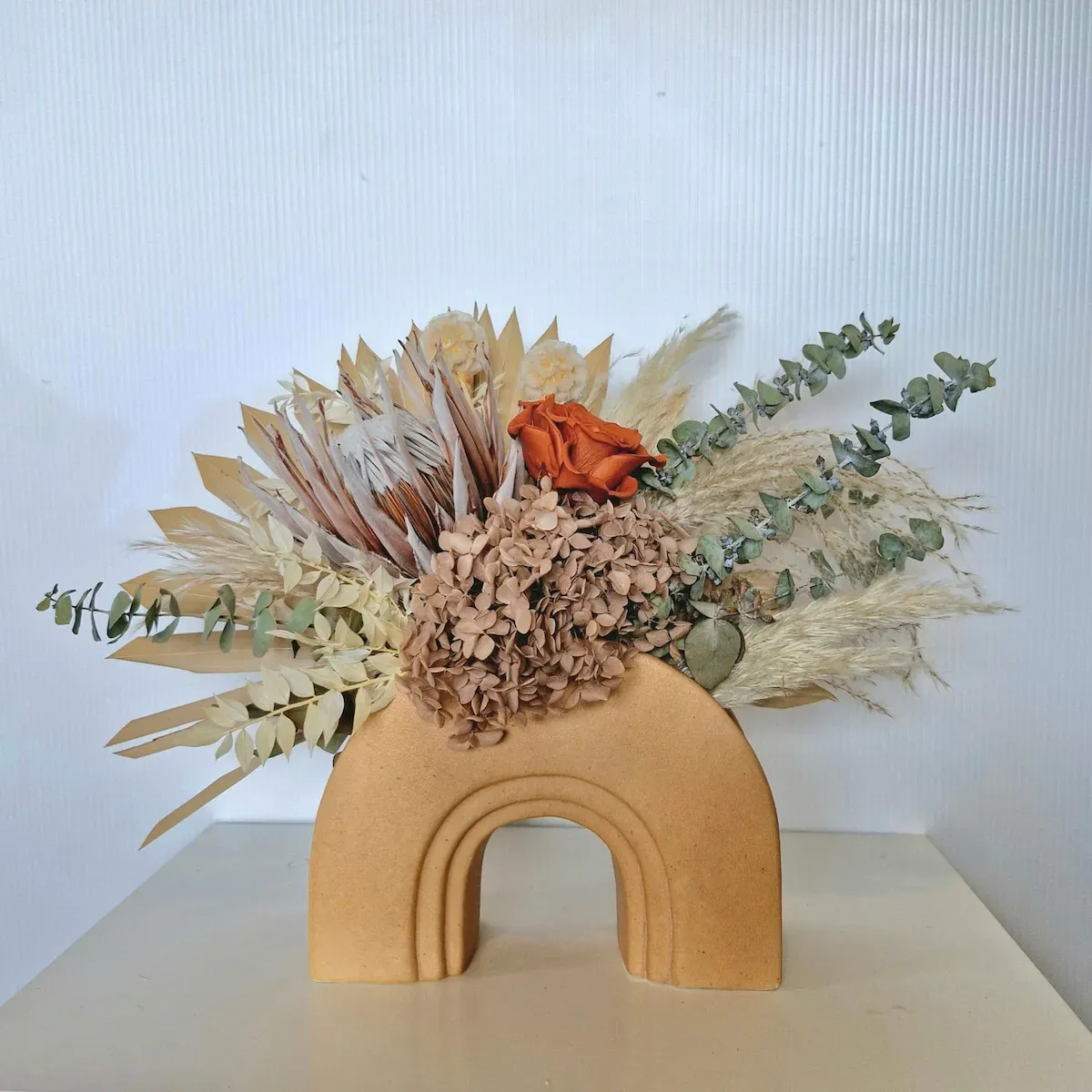 Glimmer dried floral arrangement in ceramic arch vase, featuring burnt-orange rose, coffee-brown hydrangea, and king protea.