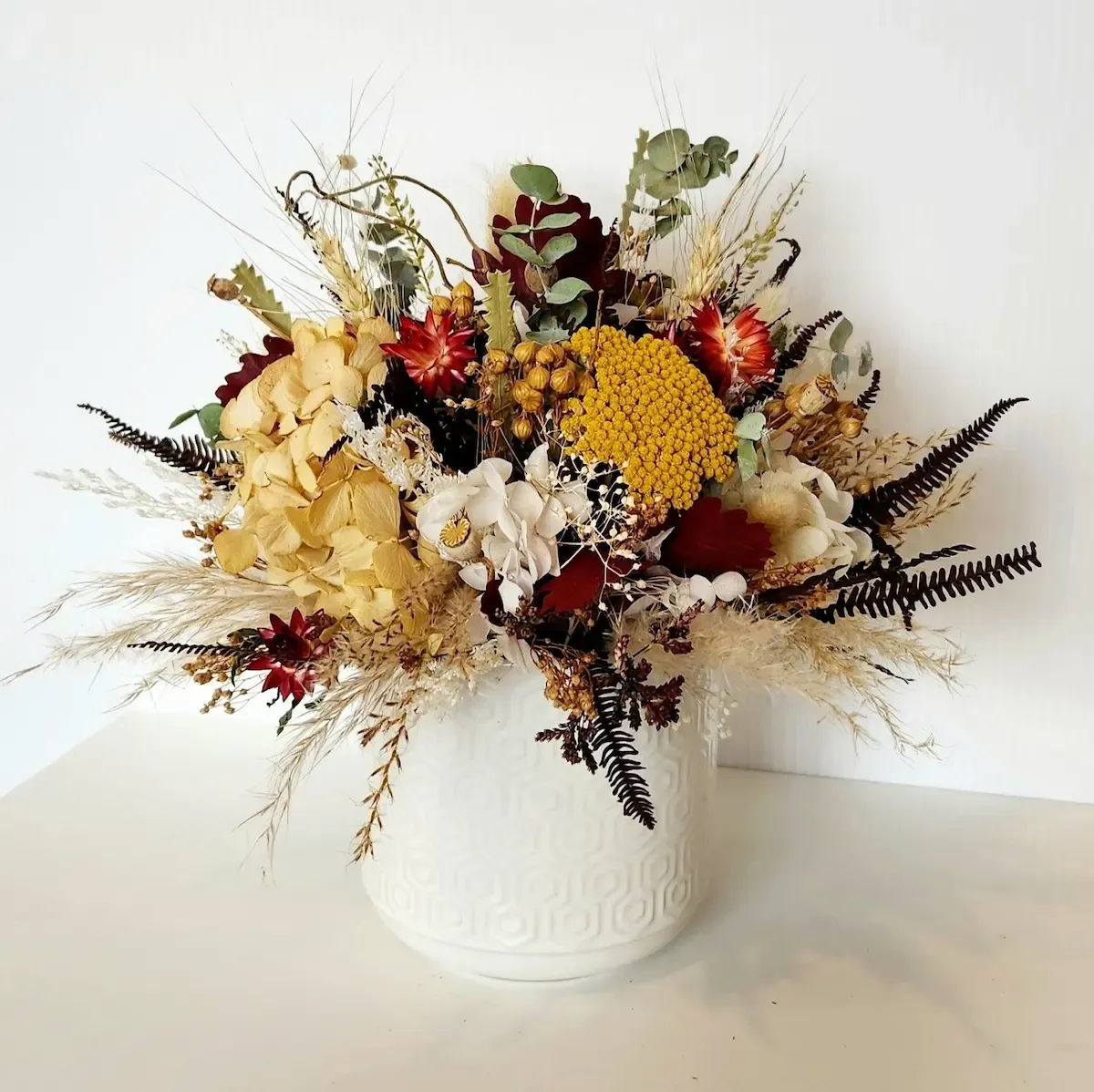 Front view of Autumn dried flower arrangement in white ceramic pot with reds, yellows, neutrals, and preserved blooms.