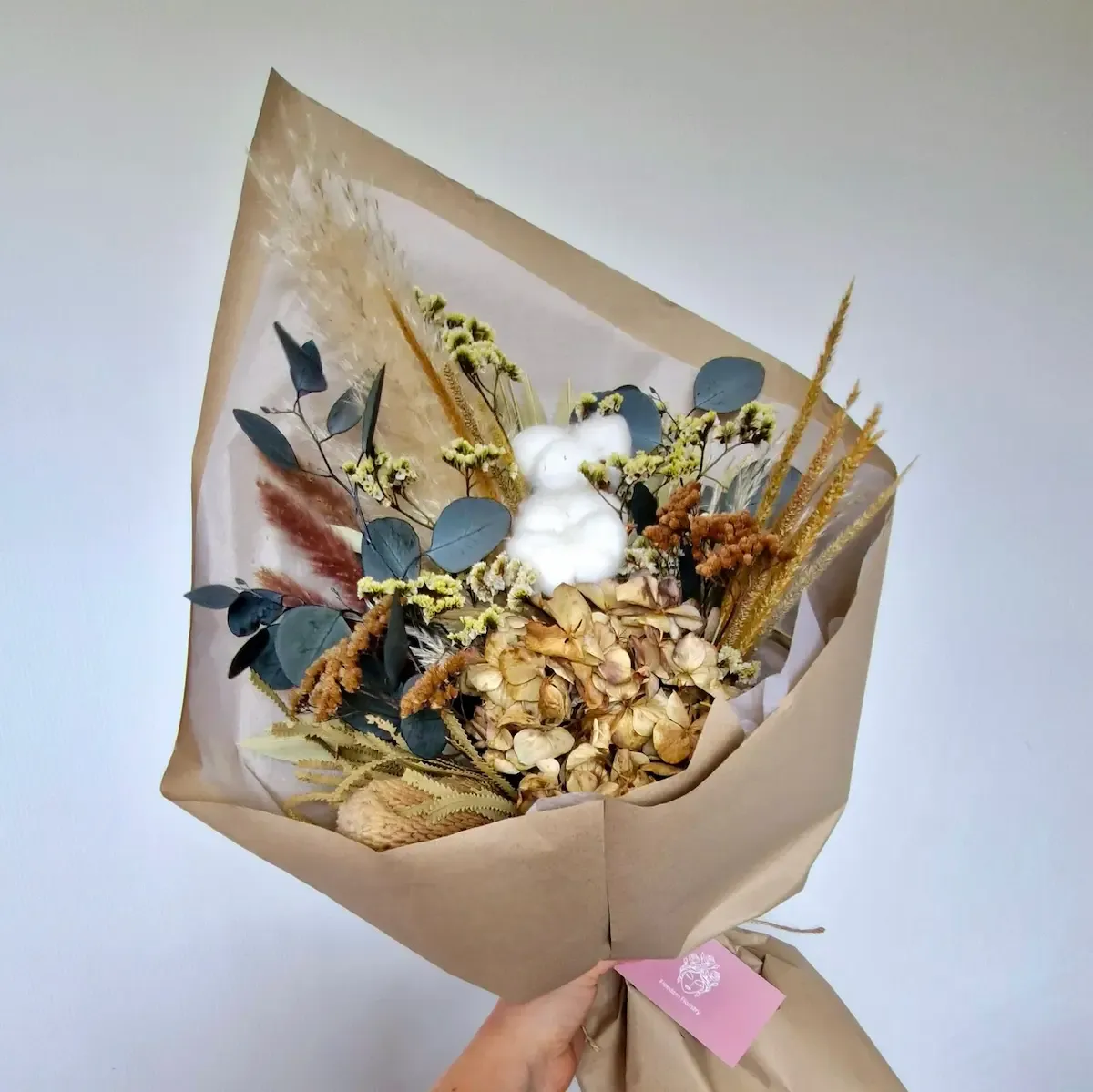 Earthy dried floral bouquet shown at an angle, featuring neutral hydrangea, banksia, cotton stems, and grasses.