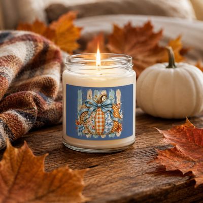 Lit patchwork pumpkin soy candle with blue label, surrounded by fall leaves, a white pumpkin, and a plaid blanket.