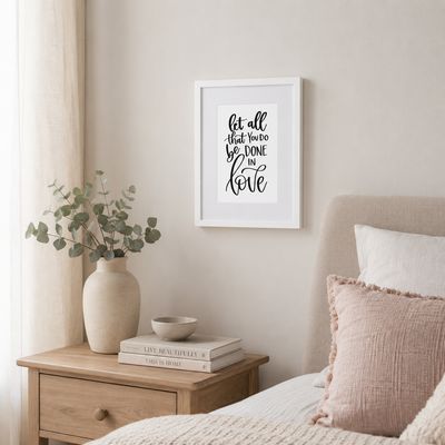 21×30 cm white framed Bible verse print, “Let all that you do be done in love,” displayed above a bedroom nightstand.