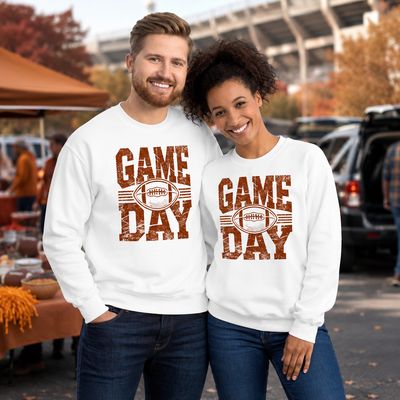 Smiling man and woman wearing matching white Game Day football sweatshirts at an autumn stadium tailgate.