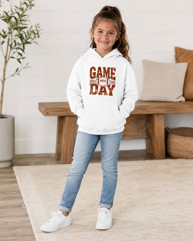 Girl wearing a white kids Game Day football hoodie with distressed rust graphic, jeans, and white sneakers.