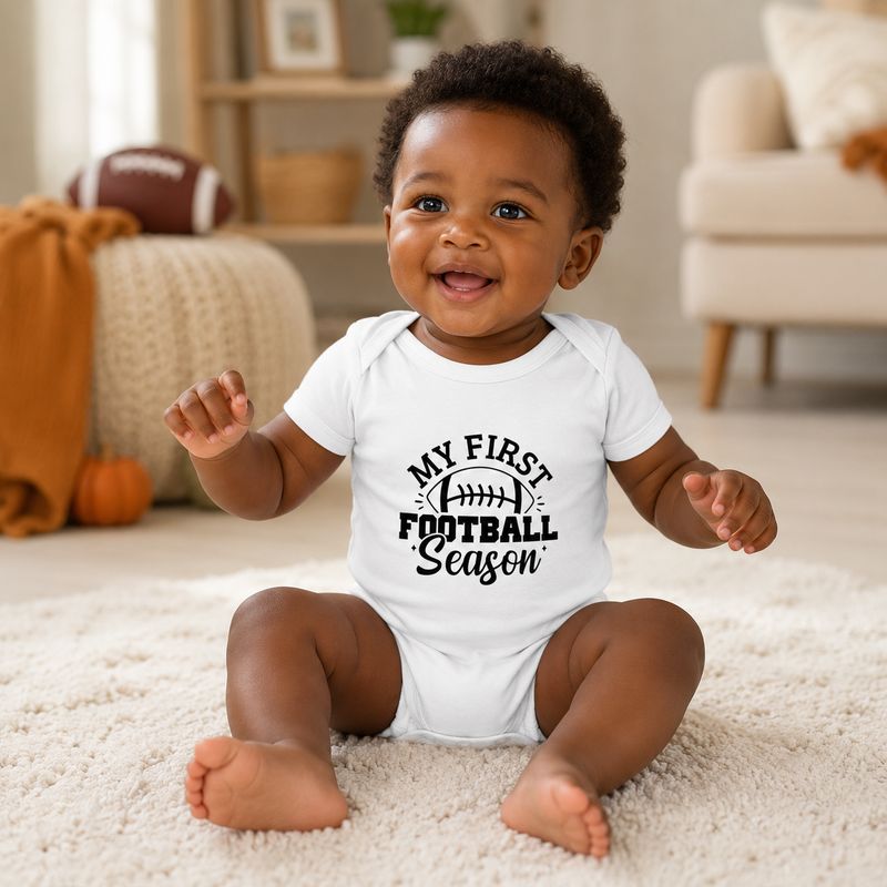 Happy Black baby in a white My First Football Season bodysuit seated in a cozy football-themed room.