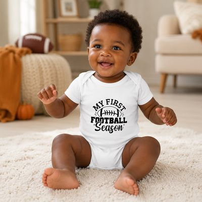 Happy Black baby in a white My First Football Season bodysuit seated in a cozy football-themed room.