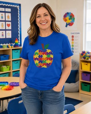 Autism Teacher Puzzle Apple Shirt, Special Education Teacher T-Shirt, Colorful Teacher Tee, SPED Classroom Shirt, Educator and Paraprofessional Gift Middle-aged teacher in a royal blue puzzle apple T-shirt inside a colorful autism classroom.