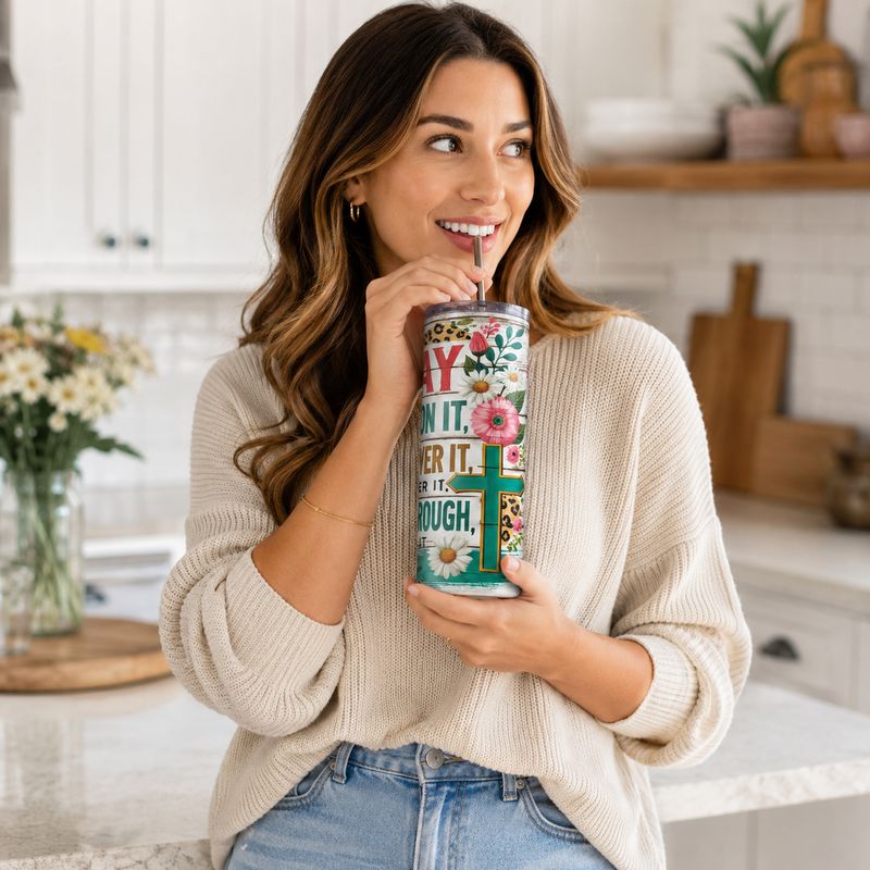 Woman sipping from a floral Christian prayer tumbler with teal cross, clear lid, and metal straw in a bright kitchen.