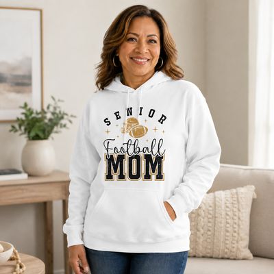 Latina woman wearing white Senior Football Mom hoodie with black and gold football design in a cozy home