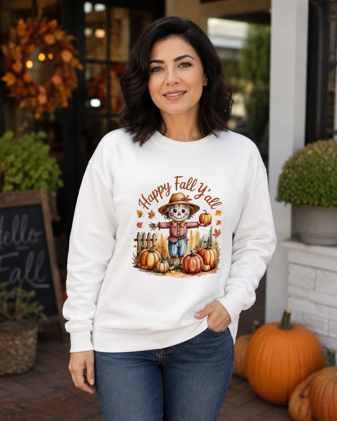 White: Woman wearing a white Happy Fall Y’all scarecrow sweatshirt on a welcoming porch decorated with pumpkins and fall foli