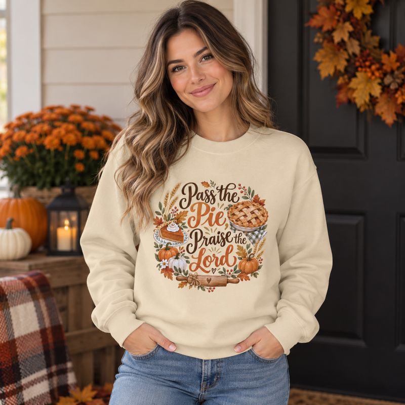 Woman wearing sand Pass the Pie Praise the Lord Thanksgiving sweatshirt on a cozy fall front porch with pumpkins and autumn d