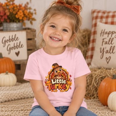 Smiling toddler girl wearing a pink Little Turkey Thanksgiving T-shirt in an autumn setting with pumpkins and fall decor.