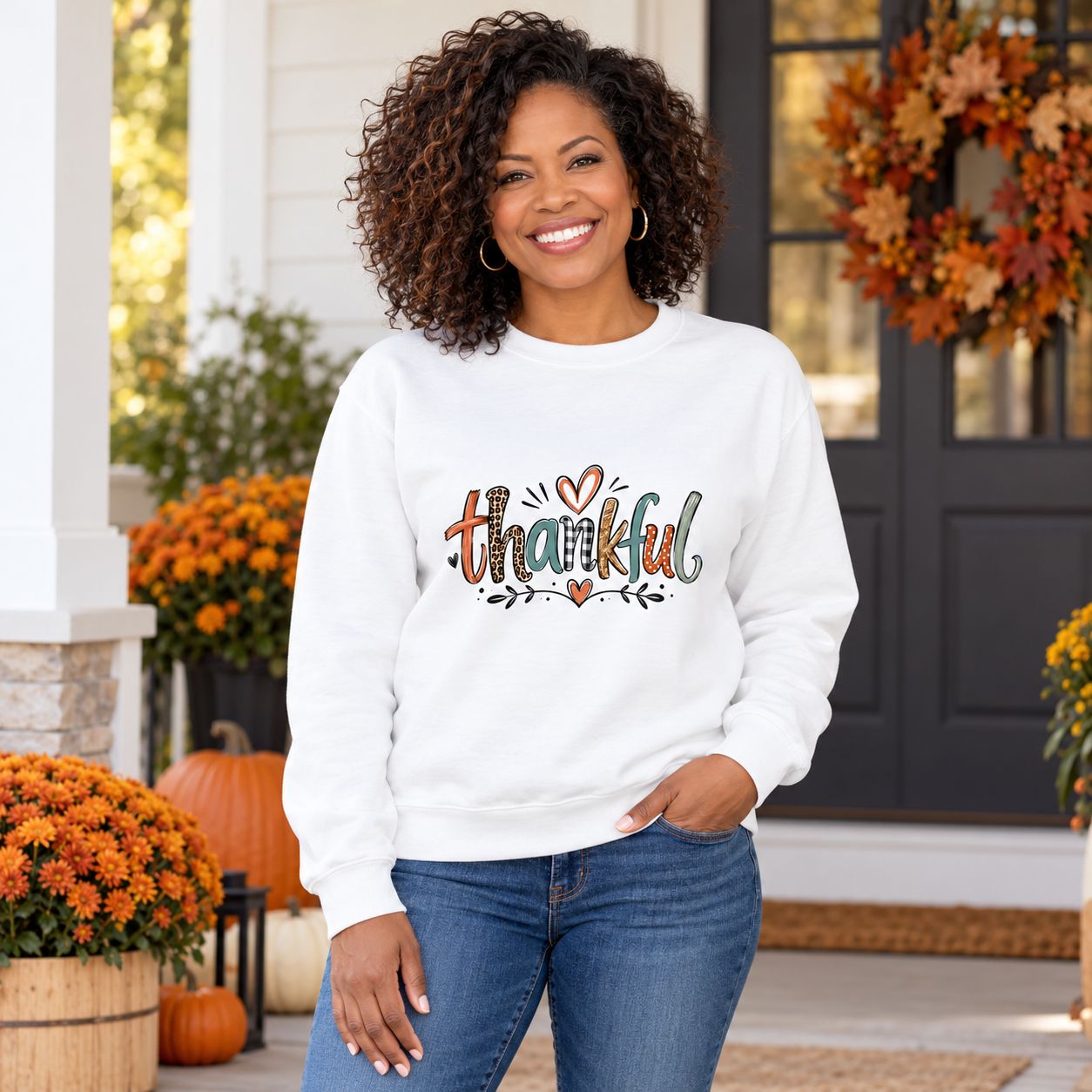 Smiling woman wearing white Thankful Thanksgiving graphic sweatshirt with colorful lettering, standing on a porch decorated w