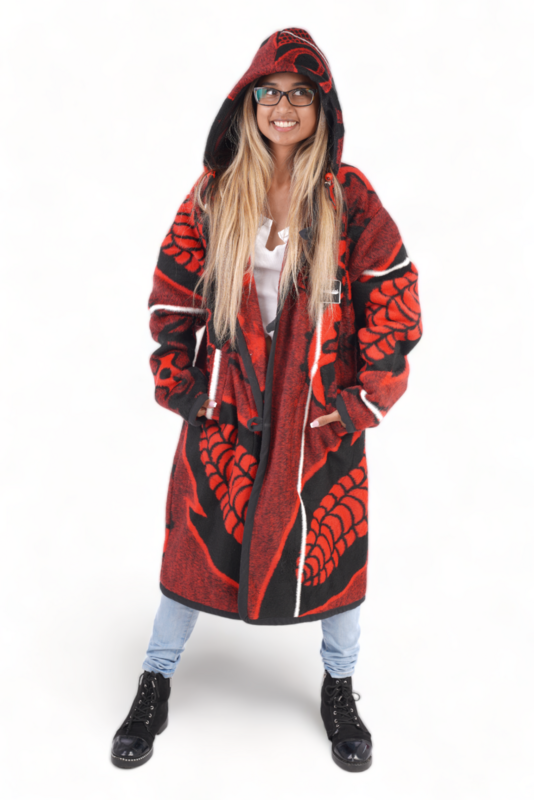 A woman in a red and black patterned Basotho blanket jacket with a hood stands confidently. She wears a white top and blue jeans.