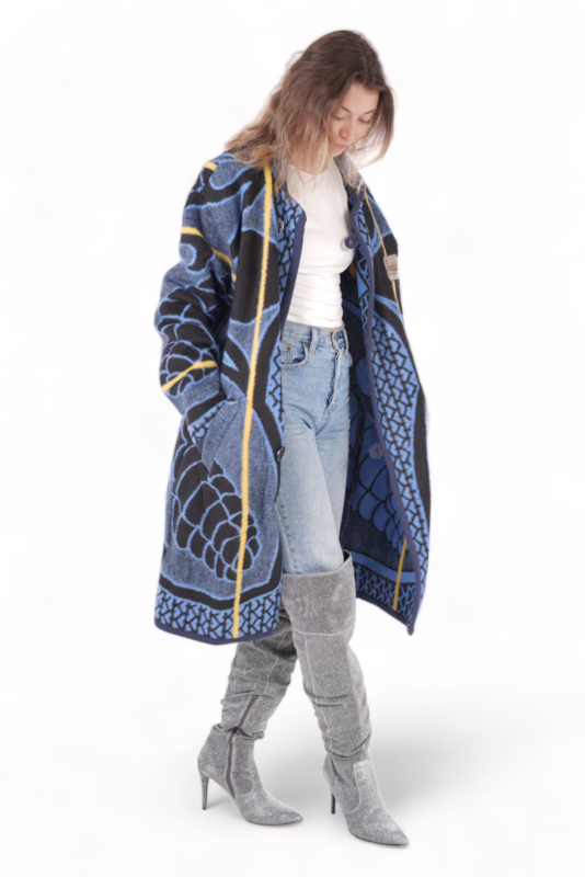 Female model in a blue patterned Basotho blanket jacket with yellow accents, hand on head, paired with shiny gray knee-high boots. She stands against a white background.