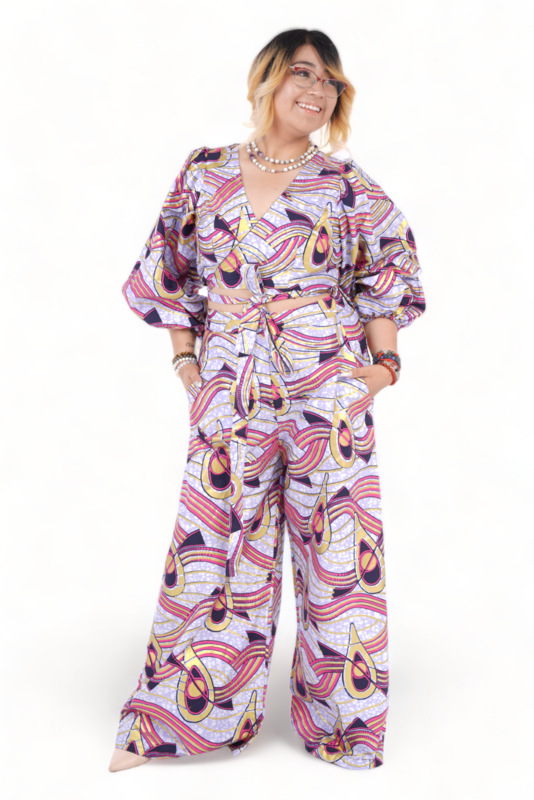 A person in a vibrant, patterned jumpsuit featuring swirling shapes in purple, pink, and yellow hues. They have a confident pose, hands in pockets.