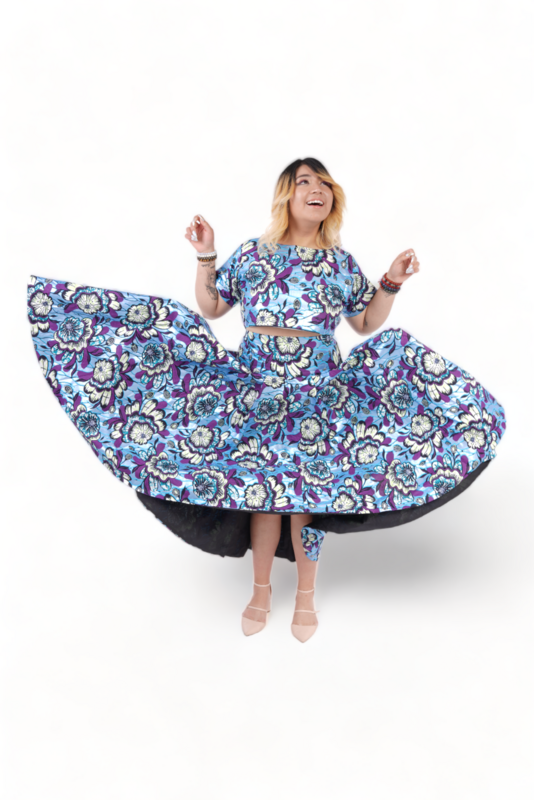 Woman wearing a vibrant floral-patterned two-piece outfit with a wide, flowing skirt. She poses cheerfully against a white background, exuding joy.