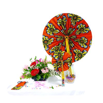 Vibrant floral arrangement with pink and red flowers in a black vase on a white table. Beside it are colorful African fans and scattered beads.