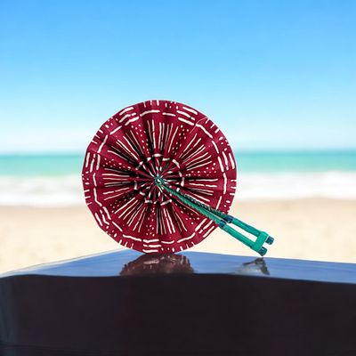 A vibrant, circular hand fan with red fabric decorated by white, symmetrical geometric patterns. The green handle is folded, adding a contrasting element.
