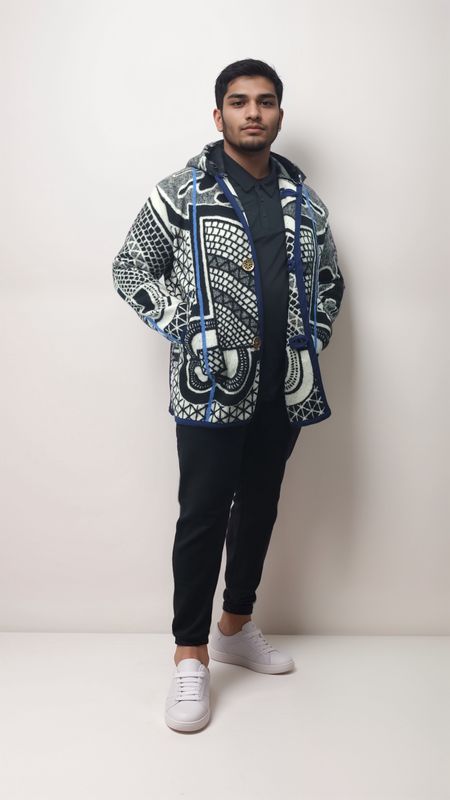 Man in a patterned jacket with geometric designs stands confidently against a light background. He's in black pants and white sneakers, exuding a casual, stylish vibe.