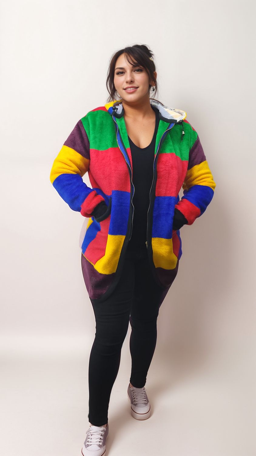 Woman in a colorful, patchwork jacket stands confidently against a plain background. She wears black pants and white sneakers, exuding a casual, joyful vibe.