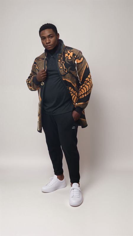 Young man in a stylish, patterned coat with orange and black accents over a black shirt and pants. He exudes confidence, standing on a plain backdrop.
