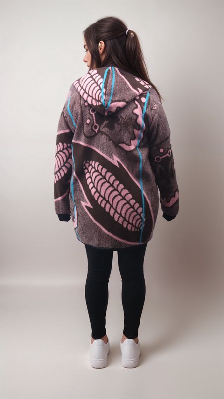 A woman stands facing away, wearing a brown hoodie with pink and blue abstract design, black leggings, and white sneakers, set against a plain backdrop.
