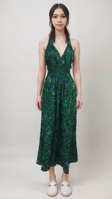A woman standing against a plain background, wearing a sleeveless, green and blue patterned dress. She exudes a calm demeanor and is wearing white shoes.