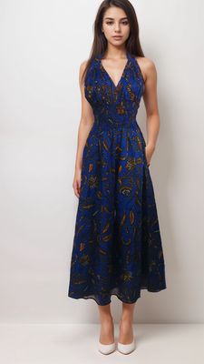 A woman in a deep blue halter dress with intricate orange patterns stands against a plain background. She has long hair, hands in pockets, and wears white heels.