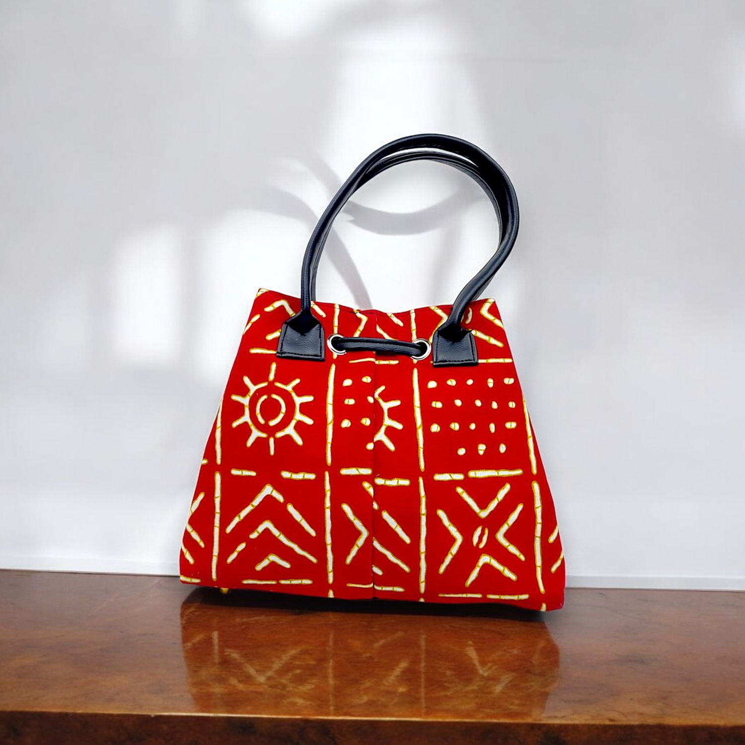 Red handbag with intricate yellow tribal patterns, black leather handles, and a drawstring closure. The design feels vibrant and artistic.