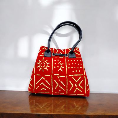 Red handbag with intricate yellow tribal patterns, black leather handles, and a drawstring closure. The design feels vibrant and artistic.