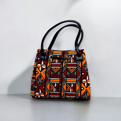 A vibrant handbag with black handles, featuring geometric patterns in orange, red, and black, conveying a bold and ethnic aesthetic.