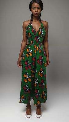 A woman stands confidently wearing a vibrant green dress with red, yellow, and black floral patterns. She pairs it with white heels, exuding elegance.