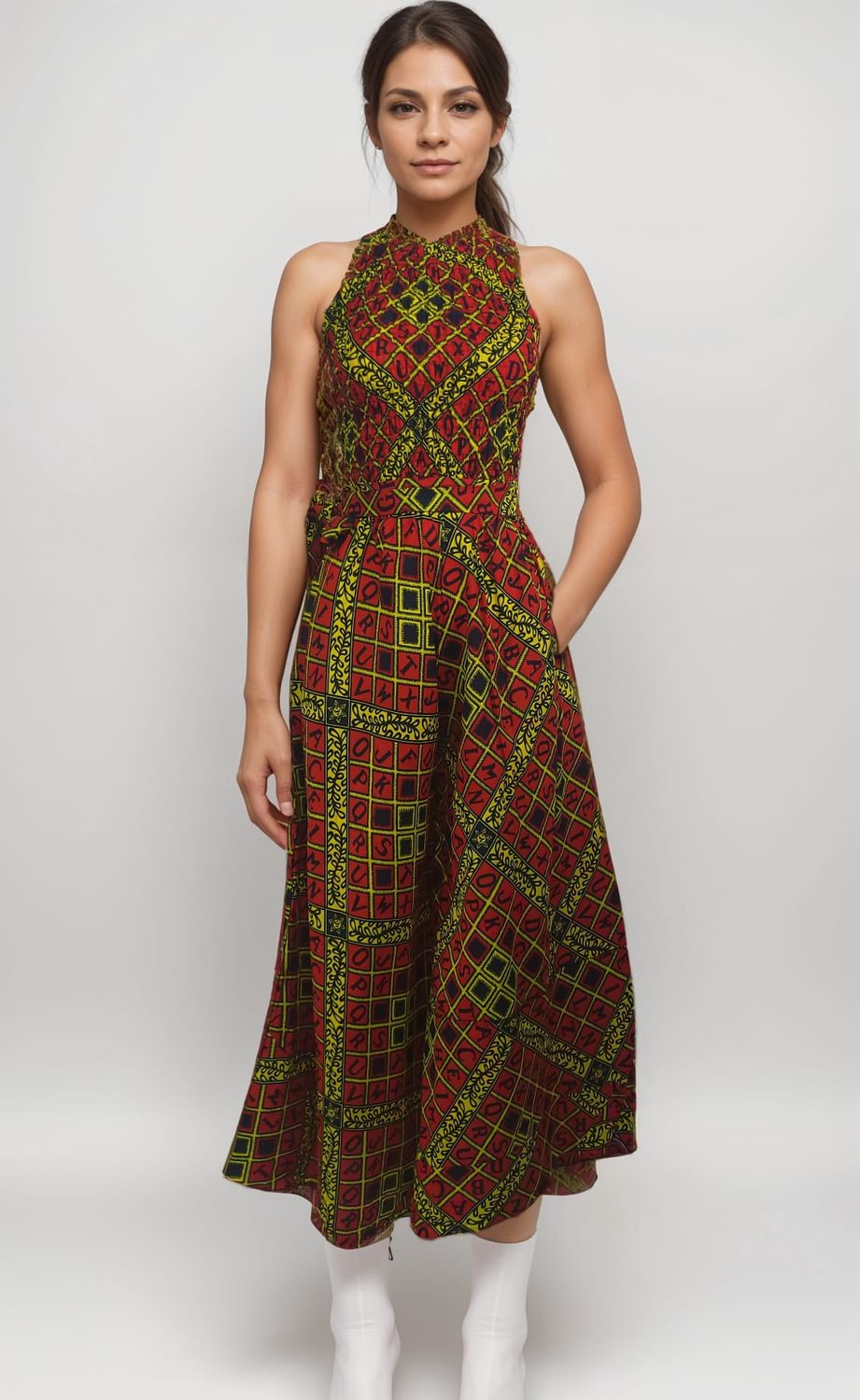 Woman in a sleeveless, patterned dress with red, yellow, and green geometric designs. She stands confidently against a plain background, wearing white boots.