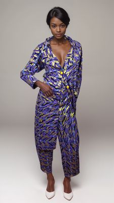 Woman in a vibrant purple and gold patterned jumpsuit with yellow buttons poses confidently against a light gray background, wearing white heels.