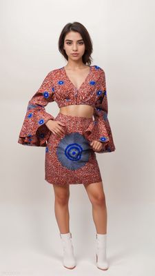 A woman with long blonde hair wears a stylish red-patterned crop top and skirt with bold blue circular designs, paired with white heels, on a neutral backdrop.