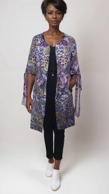 A woman stands confidently wearing a colorful, patterned kimono-style jacket over a black outfit with white ankle boots, set against a plain background.
