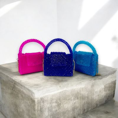 Three colorful, beaded handbags in pink, blue, and teal are artfully displayed on a concrete pedestal against a minimalist white background.