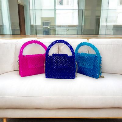 Three colorful, beaded handbags in pink, blue, and teal are artfully displayed on a concrete pedestal against a minimalist white background.