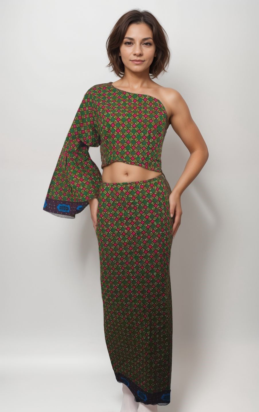A woman stands against a plain background wearing a geometric-patterned one-shoulder crop top and matching long skirt. She has an elegant, confident expression.