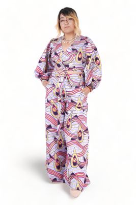 A person in a vibrant, patterned jumpsuit featuring swirling shapes in purple, pink, and yellow hues. They have a confident pose, hands in pockets.