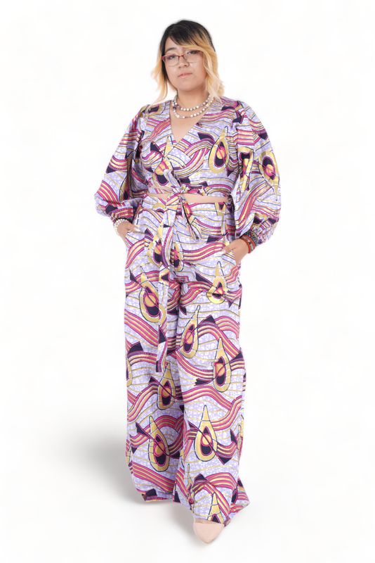 A person in a vibrant, patterned jumpsuit featuring swirling shapes in purple, pink, and yellow hues. They have a confident pose, hands in pockets.