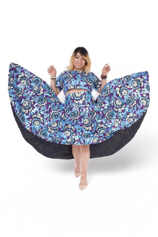 Woman wearing a vibrant floral-patterned two-piece outfit with a wide, flowing skirt. She poses cheerfully against a white background, exuding joy.