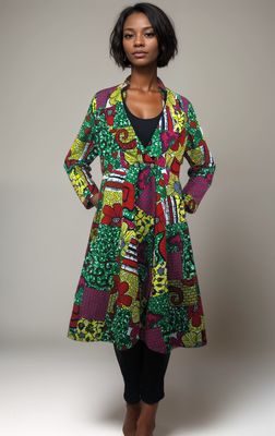 A woman models a vibrant, patterned coat with red, green, yellow, and purple designs. She wears black leggings and white heels, exuding confidence.