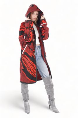 A woman in a red and black patterned Basotho blanket jacket with a hood stands confidently. She wears a white top and blue jeans.