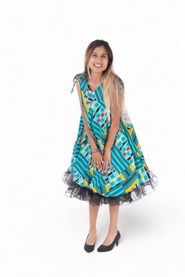 Woman smiling in a colorful, patterned dress with teal, red, and yellow accents. She is leaning slightly forward and wearing black heels. Bright and cheerful mood.