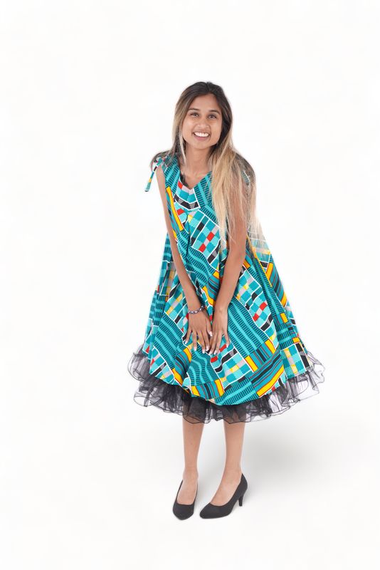 Woman smiling in a colorful, patterned dress with teal, red, and yellow accents. She is leaning slightly forward and wearing black heels. Bright and cheerful mood.